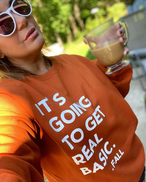 pumpkin orange sweatshirt with text screen print that says its going to get real basic for fall, women in glasses holding a clear glass of coffee