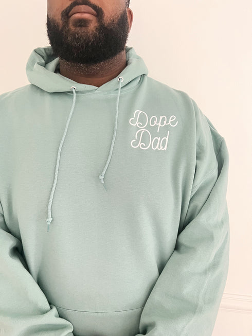 black dad behind a white background modeling an ocean blue color hoodie with screen print design that says dope dad, cute mens sweatshirt hoodie