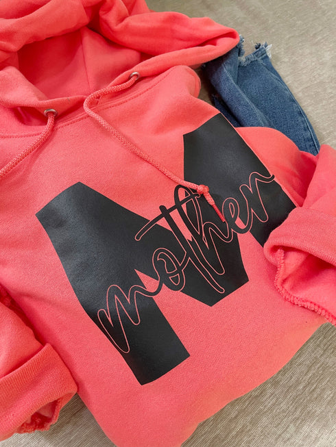 flat lay coral hoodie with black ink that says mother with a varsity letter M laying on top of skinny blue jeans for a cute cozy outfit for mothers