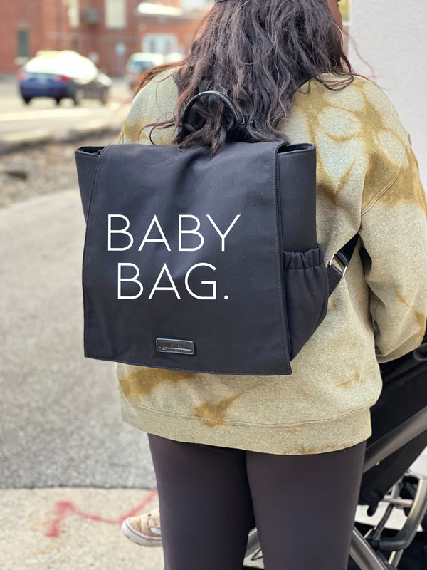 mom carrying black backpack used for a diaper bag to carry all baby items and change of clothes, the screen print on the bag says baby bag design